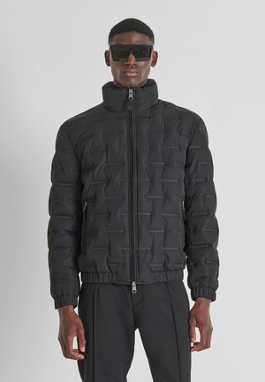 Chaqueta de plumas negra con un cuello alto, patrón texturizado, cremallera frontal y puños elásticos. Llevada sobre pantalones negros, el modelo también usa gafas de sol oscuras.