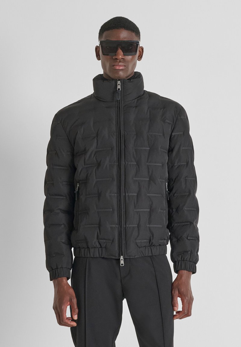 Crna puf jakna s visokim ovratnikom, teksturiranom uzorkom, prednjim patentnim zatvaračem i elastičnim manžetama. Nosila ju je preko crnih hlača, a model također nosi tamne sunčane naočale.