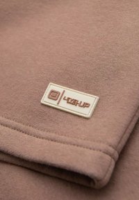 Tessuto marrone chiaro dalla texture liscia, con un'etichetta beige impressa con la scritta "LEVEL UP" in marrone. Cuciture visibili lungo i bordi.