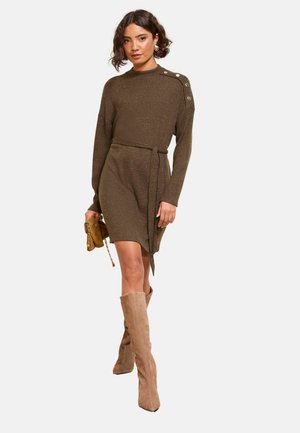 Abito a maglia a coste color verde oliva con maniche lunghe, collo alto e bottoni laterali; dettaglio in vita legato. La modella indossa stivali in suede beige fino al ginocchio.