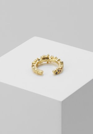 Bague texturée ouverte en or avec de petites pierres précieuses bleues placée sur un socle blanc sur fond gris.