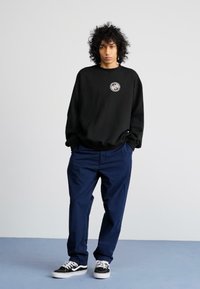 Sudadera negra con un logo en el pecho, combinada con pantalones azul marino y zapatillas negras. El fondo presenta un color claro liso.