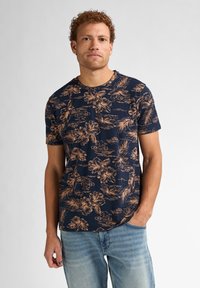 Camiseta de color marino con un patrón floral en beige. Mangas cortas, escote redondo y un corte relajado. Combinada con jeans de color azul claro.