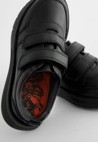 Chaussures en cuir noir avec deux bandes Velcro, bout rond et semelle texturée. L'intérieur présente une semelle intérieure rouge avec un motif graphique.