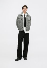 Chaqueta de mezclilla gris con un patrón sutil, dos bolsillos en el pecho y un logo. Combinada con una camisa blanca, una corbata negra y pantalones de pierna ancha negros.