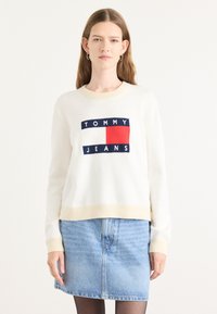 Hvit strikkegenser med beige detaljer, med et marineblått og rødt logodesign som sier "TOMMY JEANS." Båret med et lyseblått denimskjørt.