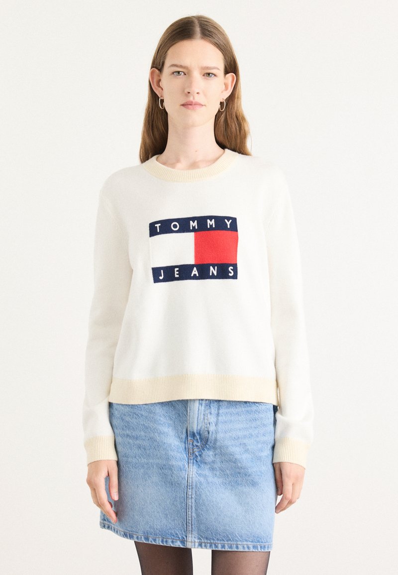 Hvit strikkegenser med beige detaljer, med et marineblått og rødt logodesign som sier "TOMMY JEANS." Båret med et lyseblått denimskjørt.