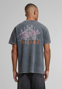 Pilkas medvilninis marškinėliai su grafika, vaizduojančia Duomo di Milano baltos ir rožinės spalvos, po ja spausdintas užrašas "Milano" oranžine spalva. Trumpomis rankovėmis, laisvo kirpimo.