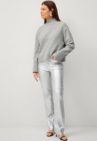 Maglione grigio a maglia con collo alto e polsini a coste, abbinato a pantaloni a zampa d'elefante argentati e lucenti. Modella in sandali neri con cinturini.