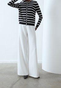 Černobílý proužkovaný kardigan s knoflíky, kombinovaný s širokými bílými kalhotami a tmavými botami, na neutrálním pozadí.