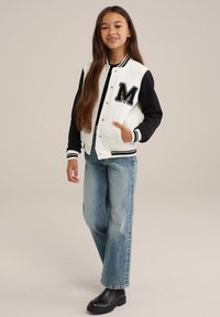 Veste varsity noire et blanche avec un grand patch "M", associée à un jean droit bleu clair et des bottines noires.