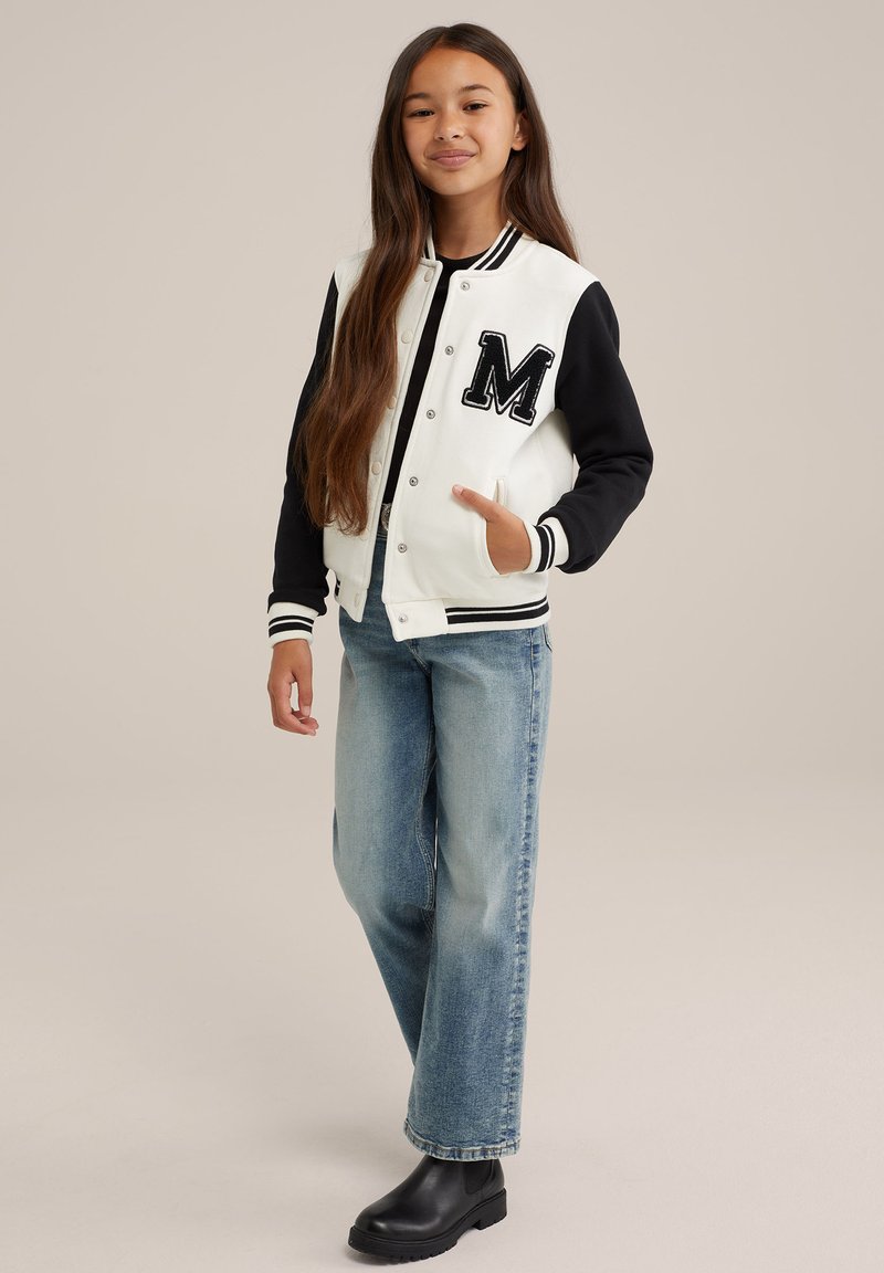 Veste varsity noire et blanche avec un grand patch "M", associée à un jean droit bleu clair et des bottines noires.