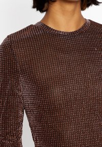 Brun, strukturerad topp med ett rutig mönster och en lätt glans. Har en rund halsringning och långa ärmar.