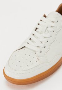 Baskets en cuir blanc avec un bout perforé, lacets blancs plats et semelle en caoutchouc marron. Présente une texture lisse et des détails de couture subtils.