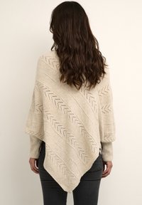 Beige knitted poncho with an asymmetric hem, featuring a textured zigzag pattern and ribbed sleeves, worn over dark jeans.