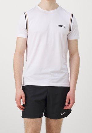 Mężczyzna w białej koszulce BOSS z czarnymi i beżowymi paskami na rękawach oraz czarnych szortach Nike na jasnym, jednolitym tle.