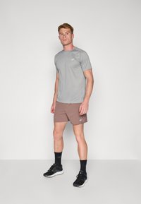 Graues kurzärmliges Sportshirt, braune Shorts, schwarze Laufschuhe, stehend auf einer grauen Fläche. New Balance-Logo auf den Kleidungsstücken. Einfaches Design.