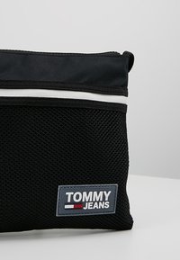 Borsa a rete nera con texture intrecciata, dotata di una striscia bianca e di un patch rettangolare con il logo "TOMMY JEANS" in blu e bianco.