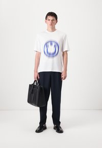 T-shirt blanche à manches courtes avec un logo graphique bleu, associé à un pantalon sombre et un sac fourre-tout en cuir noir avec deux poignées supérieures.