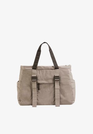 Beige tote bag with dark handles, front zipper pocket, side pockets, and two adjustable buckled straps on the front.