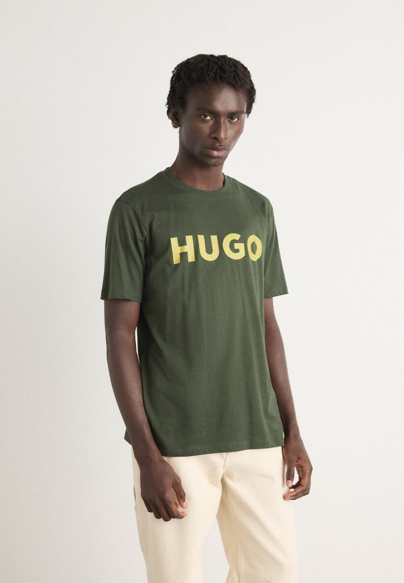 Zöld pamut póló merész sárga "HUGO" felirattal az elején; rövid ujjú, kerek nyakú, laza szabású. A modellen krém színű nadrág van.