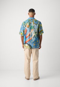 Camisa azul de manga corta con estampados vibrantes de frutas en naranja y verde, manos y estrellas decorativas. Combinada con pantalones cargo beige.