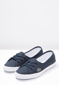 Chaussures en toile bleu marine avec semelles en caoutchouc blanches, bout rond, design à lacets, et un petit logo de crocodile vert sur le côté.