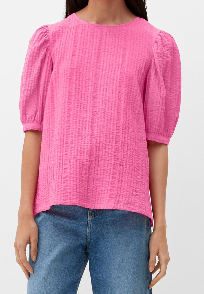 Femme portant un haut rose vif texturé avec des manches bouffantes à mi-bras et un jean en denim bleu, montré du cou aux hanches.