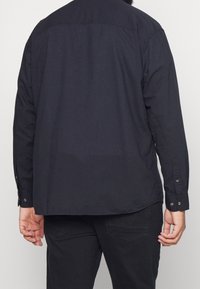 Camicia a maniche lunghe di colore blu scuro, realizzata in cotone. Presenta un polsino con bottone e un colletto classico, con orlo dritto e senza un motivo visibile.