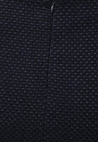 Gros plan d’un tissu noir texturé avec un petit motif carré et une fermeture éclair noire partiellement visible en haut au centre.