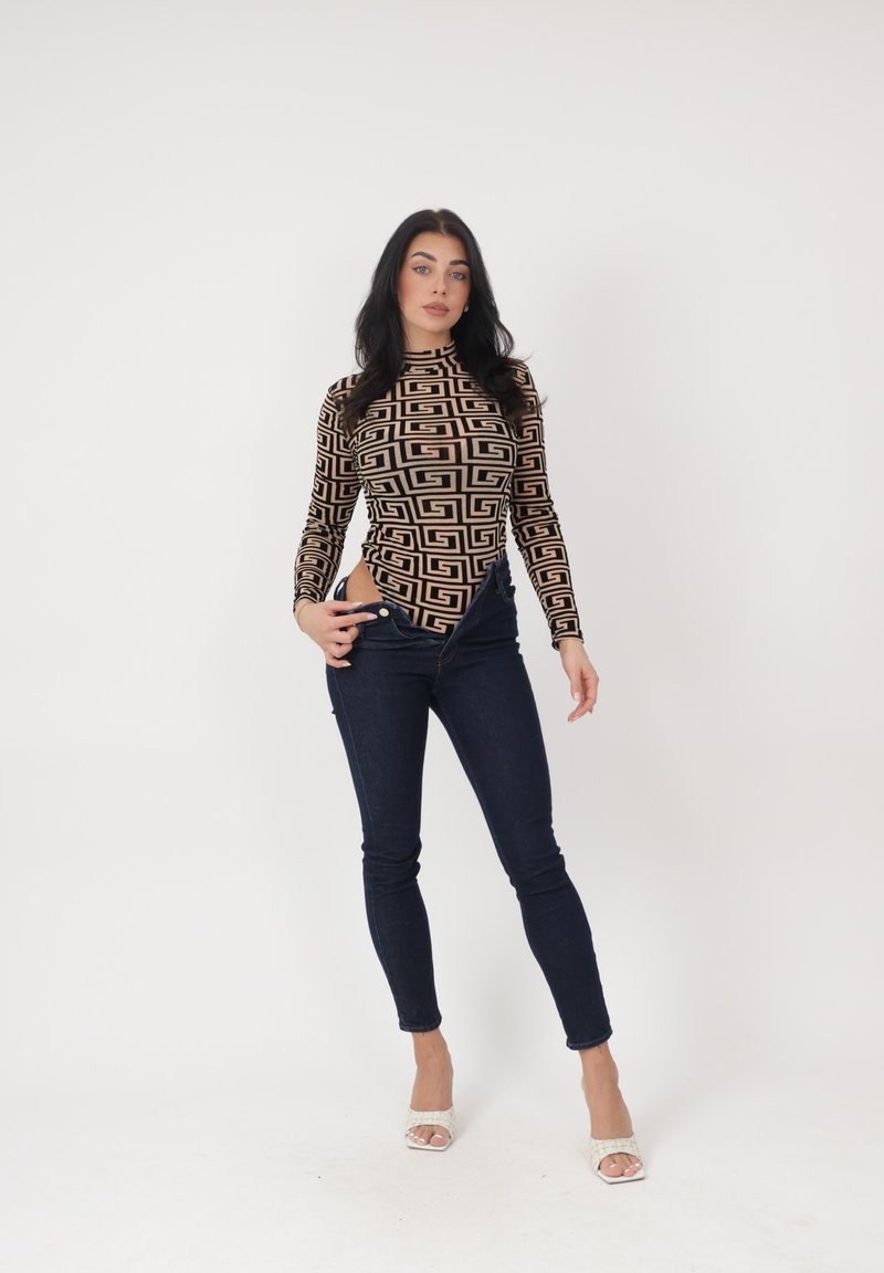 Mujer de pie con blusa de manga larga estampada, vaqueros oscuros desabrochados y tacones blancos abiertos, sobre fondo blanco liso.