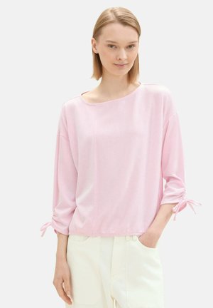 Top lungo a maniche lunghe di un rosa chiaro a coste, con scollo a barca, maniche arricciate con fiocchi, abbinato a pantaloni bianchi. Texture liscia, vestibilità comoda.