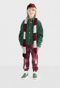 Barn ler, bär grön manchesterskjorta, röd- och vitrandig halsduk, röda byxor med stjärnmönster, gröna slip-on skor med prickar och röd keps.