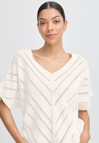 Sweter na drutach w rozmiarze oversize w kolorze kremowym, z dekoltem w kształcie litery V oraz wzorem w zygzak z elementami ażurowymi. Krótkie rękawy i luźny fason.