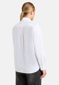 Camicia bianca con colletto classico, maniche lunghe e dettaglio a pieghe sul retro, abbinata a pantaloni grigio scuro. Tessuto dalla texture liscia.