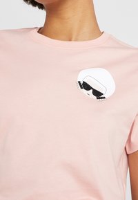 Světle růžové tričko z měkké bavlny s grafikou postavy v brýlích. Kulatý výstřih, krátké rukávy, jednoduchý design.