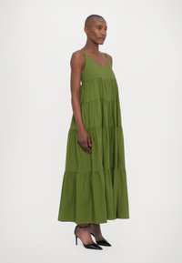 Vestito maxi verde con un design a strati, realizzato in un tessuto leggero. Caratterizzato da spalline sottili e una silhouette fluida. Indossato con tacchi neri.
