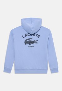 Vaaleansininen huppari, jossa on kiristysnaru ja musta Lacoste-logo sekä krokotiiligravyyri takana. Pehmeä puuvillasekoitekangas.