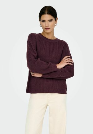 Pull en maille texturée violet profond avec un col rond et des manches oversized. Associé à un pantalon de couleur claire, créant un contraste.