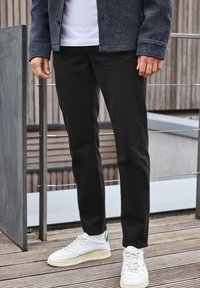Pantaloni neri slim fit, realizzati in cotone; abbinati a sneakers bianche con dettagli verdi. In piedi accanto a una ringhiera di legno.