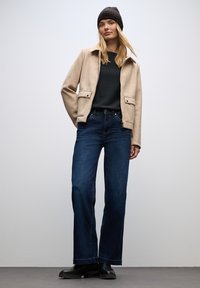 Chaqueta de mezcla de lana beige con dos bolsillos frontales, suéter verde oscuro debajo y jeans anchos azules; unas botas negras hasta el tobillo completan el look.