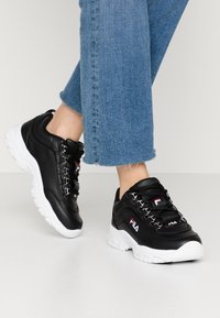 Zapatillas sintéticas negras con suelas de goma blanca, lados texturizados y ojales metálicos. Presenta acentos de logo en rojo y blanco en el lateral.