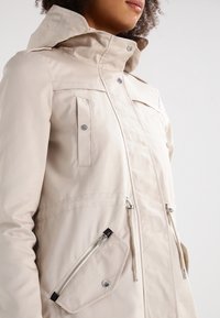 Vaaleanbeige vedenpitävä takki, jossa on korkea kaulus, useita taskuja sekä napin ja vetoketjun yhdistelmä sulkemisessa. Sileä pinta ja hienovaraiset ompeleet.