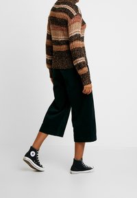 Personne marchant vêtue d'un pull en tricot rayé multicolore, de pantalons cropped noirs à jambes larges et de baskets montantes noires sur un fond blanc uni.