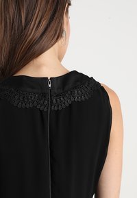 Žena v bezramínkových černých šatech s krajkovým límcem a zadním zipem, viditelný pouze její zadní část, část vlasů a paže.