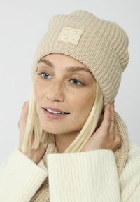 Beige gebreide beanie met een geribbeld textuur en een vierkante logo patch. Vergezeld door een beige sjaal, casual gedragen.
