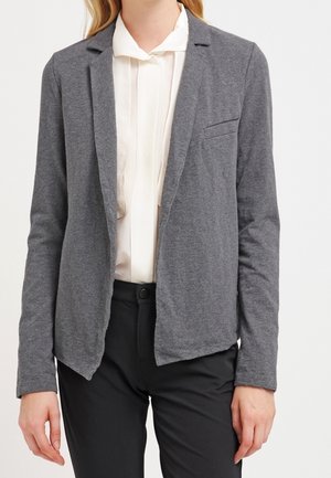 Blazer gris en tissu doux et texturé, avec une poche poitrine unique et un design ouvert, porté sur un chemisier de couleur claire.