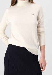 Světlý krémový rolák z měkké pleteniny, se skrytým logem na hrudi a pružnými manžetami, v kombinaci s tmavě proužkovanou sukní.