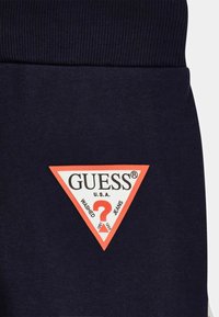 Dunkelblaues Gewebe mit einem gerippten Taillenbund. Enthält ein markantes dreieckiges Logo in Weiß und Orange mit dem Text "GUESS® U.S.A. WASHED JEANS."