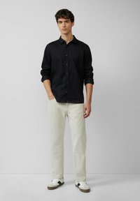 Chemise en lin noire avec fermeture à boutons, manches longues et poignets retroussés, associée à un pantalon en denim de couleur claire et des baskets décontractées.